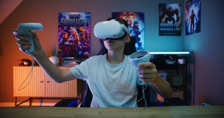 Young gamer in VR headset plays virtual reality online video game using wireless controllers. Boy...