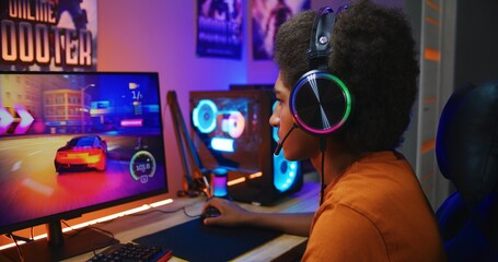 African American guy, teenage gamer in headphones plays in online 3D race game on modern powerful...
