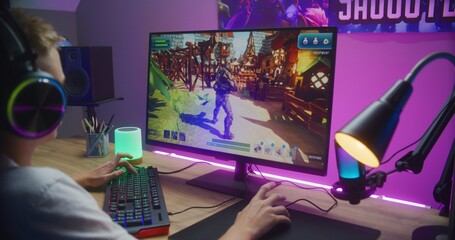 Young gamer plays third person shooter on personal computer. Schoolboy in headphones enjoys online video game at home. Desk illuminated by RGB LED strip light. Concept of gaming at home. Back view.