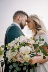 Happy bride and groom are hugging at the wedding day in the garden. Bride holding the beautiful and luxurious bouquet of fresh flowers and eucalyptus.