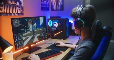 Young gamer plays in first person shooter on PC at home. Computer monitor with displayed online video game live stream or cybersport championship. Desk illuminated by RGB LED strip light.