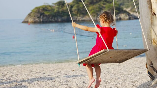Little girl on wooden swing on Oludeniz beach. Summer holiday in Oludeniz, Turkey. Girl ride on a swing on sea coast, clear water. Travel concept