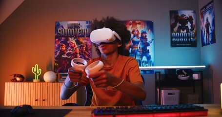 Teenage gamer in VR headset plays virtual online video game using wireless controllers. African American guy having fun in leisure time in his room. Gaming at home. View from PC screen perspective.