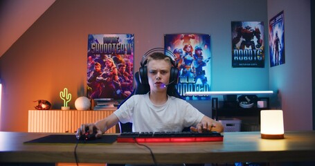 Young gamer in headphones speaks with teammates, plays online video game on computer in cyber gaming neon room. Excited boy enjoys multiplayer cybersport competition. POV from PC screen perspective. © Framestock