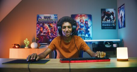 Professional African American teenage gamer in headphones speaks into mic, plays online video game...