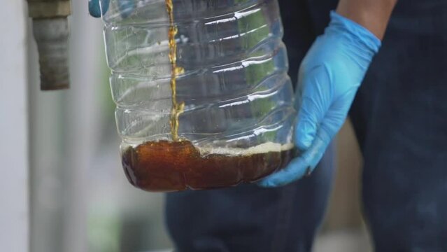 hand of man in blue rubber gloves opens a tap filling refined biodiesel into a clear plastic water tank.