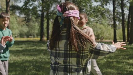 Blindfolded girl chasing friends running around in park while playing blind mans buff outdoors on summer day