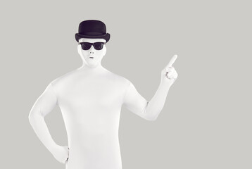 Disguised man dressed in white bodysuit pointing to side with forefinger. Portrait of faceless man wearing full body spandex costume, black bowler hat and sunglasses pointing finger to copy space