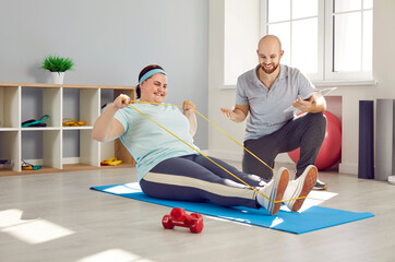 Friendly male nurse, trainer or health care worker helping fat woman doing stretching exercises with rubber band sitting on the floor. Man physiotherapist helping overweight woman in rehab.