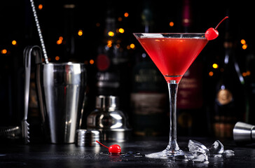 Manhattan alcoholic cocktail drink with bourbon, red vemuth, bitter, ice and cocktail cherry in glass, dark bar counter background, bar tools and bottles