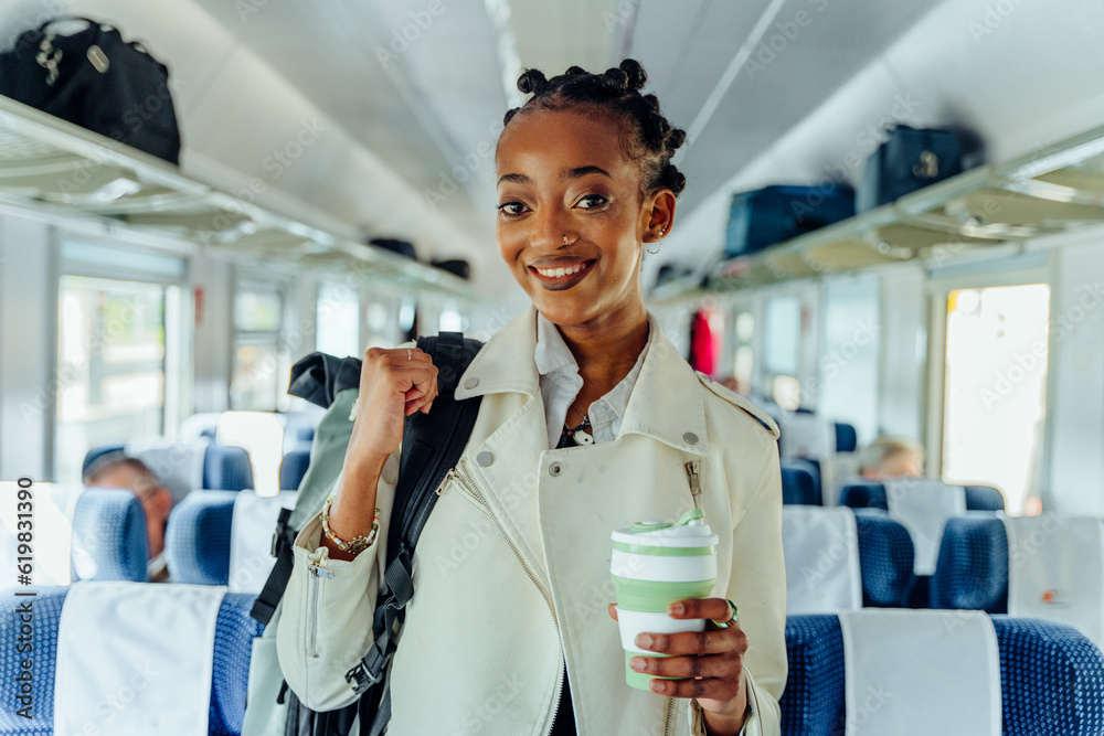 Wall mural Business trip travel long short distance public transport concept. Portrait of african female traveler In white jacket with backpack smiling drinking coffee on train indoor.