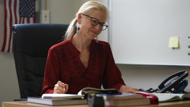 Closeup of tired woman teacher with frustration at school classroom desk with US American flag in the background.