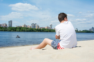 On a sunny summer day, a boy sits on the sand near the river.