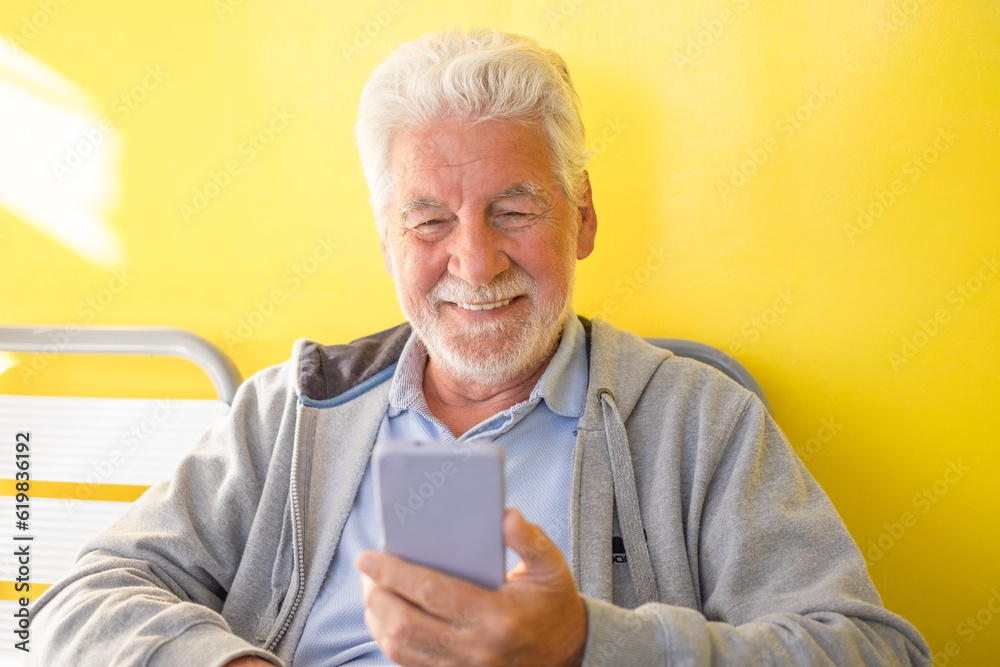 Sticker Smiling relaxed senior white haired man sitting on yellow background using mobile phone. Elderly modern grandfather enjoying tech and social