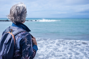 Back view portrait of senior gray haired woman in denim jacket and backpack standing in front to...