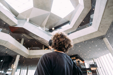 Trendy millennial hipster man with curly afro hair wear wireless sound isolating headphones. Student or solo tourist look up at ceiling of modern architecture building. Travel and explore new location