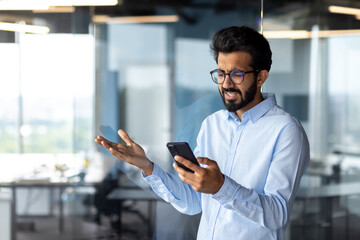 Upset and sad hispanic man inside office standing near window, businessman received message notification with bad news, man holding phone, reading information on smartphone app
