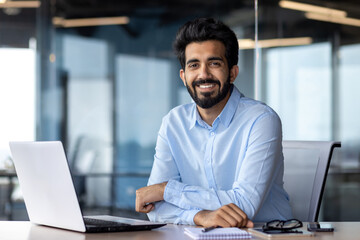 Fototapeta na wymiar Portrait of happy and successful businessman, indian man smiling and looking at camera, satisfied with achievement results man working inside office building using laptop at work
