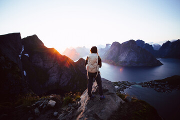 Unrecognizable female hiker stand on top of mountain summit in dark evening light. Last sunrays go over mountain range over fjord in distance. Epic hiking destination in Lofoten, Norway