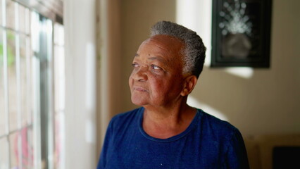 Contemplative Senior woman stands by window staring outside at home. Thoughtful pensive expression of an African American elderly person in 70s