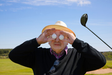 Senior man holds golf balls in front of his eyes against the background of a golf course and golf club. Concept of golf crazy , crazy about golf or golf mad.