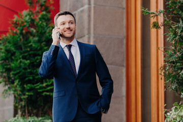 Glad entrepreneur speaks on phone, wears formal black suit, stands outdoor. Waiting for business partner near cafe/restaurant.