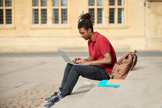 Male Student Working On Laptop Sitting On StepsOutside University Building In Oxford UK