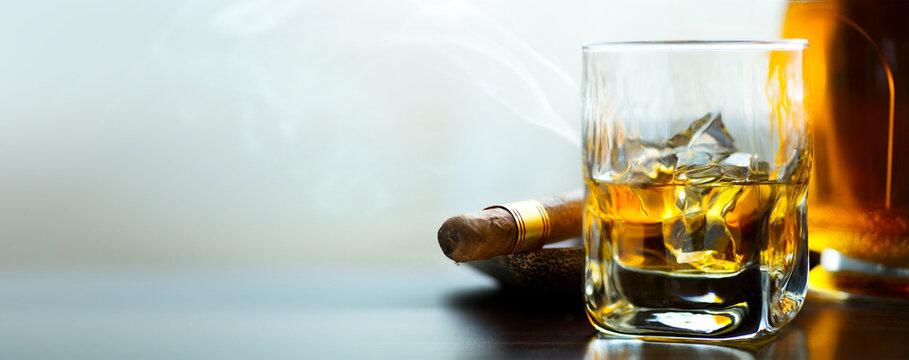 A bottle and a glass of whiskey with ice and a steaming Cuban cigar on a table against a light background. Men's club banner idea. Copy space for text
