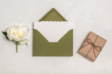 Flat lay invitation or greeting card mockup with green envelope, craft gift box and one white peony flower on light background.
