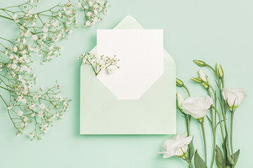 Delicate white eustoma and gypsophila flowers, green envelope and blank card on a green background. Flat composition.Greeting card, invitation.