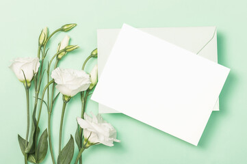 Delicate white eustoma flowers, green envelope and empty card on green background. Flat composition.Greeting card, invitation.
