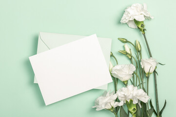 Delicate white eustoma flowers and white carnations, a green envelope and a blank card on a green background. Flat composition.Greeting card, invitation.