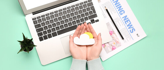 Female hands with paper cloud, laptop and newspaper on color background. Weather forecast concept
