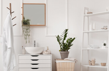 Sink bowl with bath accessories on chest of drawers, houseplant and bathrobe in bathroom