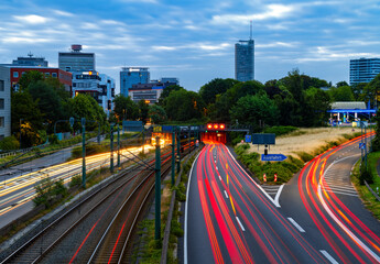Essen city with curved motorway called “Ruhrschnellweg“ in Ruhr basin. Tunnel, 4 lanes and tram rails in morning blue hour twilight with colorful light traces and skyline silhouettes of tall buildings