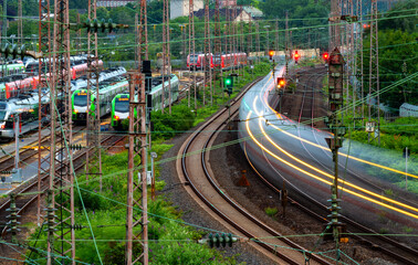 Railway depot infrastructure and main line tracks. Blue hour near station of Essen Germany with signals, power poles and passing fast train in motion. Catenary, glistening tracks at morning twilight.
