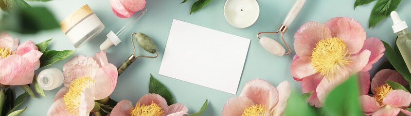 Top view of natural cosmetics and pink peonies on pastel blue background with white card for invitation, logo or design