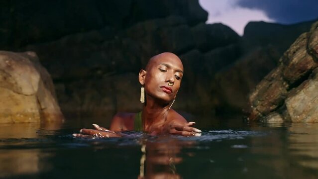 Trans sexual black fashion model poses chest deep inside natural pool surrouded by rocks on tropical island at night. Androgynous ethnic fashion model in the middle of backwater exotic scenic location