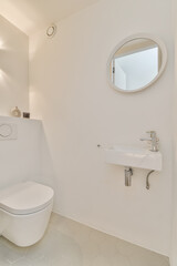 a white bathroom with a toilet and sink in front of a round mirror on the wall behind it is a small window