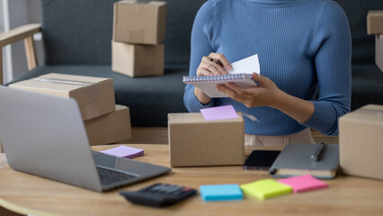 Asian woman online store business owner, online shop owner working from home, posting products for sale on website and accepting orders and packing products for customers with private courier service.