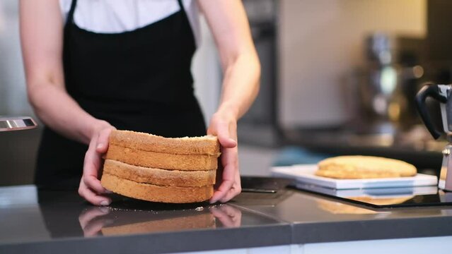 Female confectioner preparing biscuit layers custom cake for wedding celebration woman in apron smearing white sugar free cream around fresh pastry layers on stand closeup