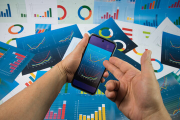 Ręka wskazująca wykres wyświetlany na telefonie komórkowym trzymanym w dłoni na tle plansz z wykresami.