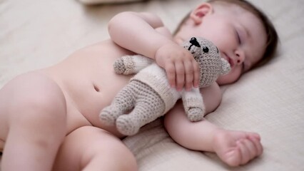 Chubby little boy with closed eyes sleeping on soft bed hugging knitted toy bear with hand adorable baby resting in nursery at home after walk on street