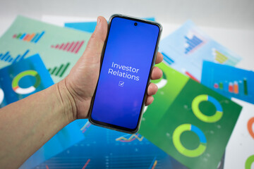 Ręka trzymająca telefon na którym jest wyświetlany napis investor relations na tle plansz z wykresami.