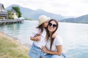Portrait of cute daughter and young mom. Outdoor photoset by the lake. Clear blue water. Happy family concept. Girl kiss mom