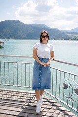 Portrait of young and stylish woman stand on bridge and smile. Background of beautiful lake. Clear blue water.