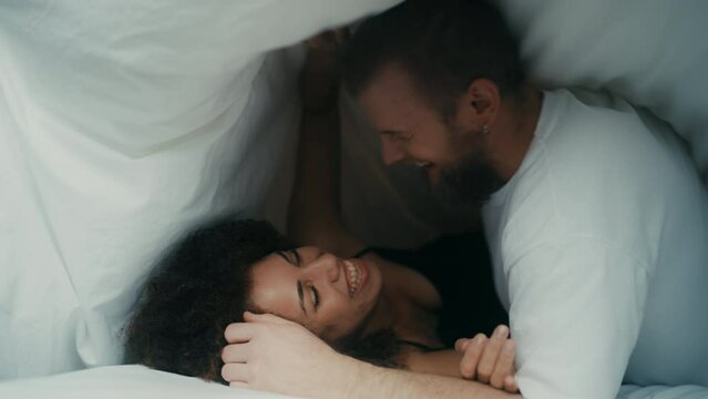 Happy young couple laughing under white linen in comfortable bed, love story