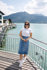 Portrait of young and stylish woman stand on bridge and smile. Background of beautiful lake. Clear blue water.