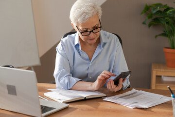 Fototapeta na wymiar Confident stylish european middle aged senior woman using smartphone at workplace. Stylish older mature 60s gray haired lady businesswoman with cell phone in office. Boss leader using internet apps