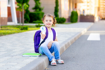 a schoolgirl child with a backpack and bills in her hands sits on the curb at the school and shows...